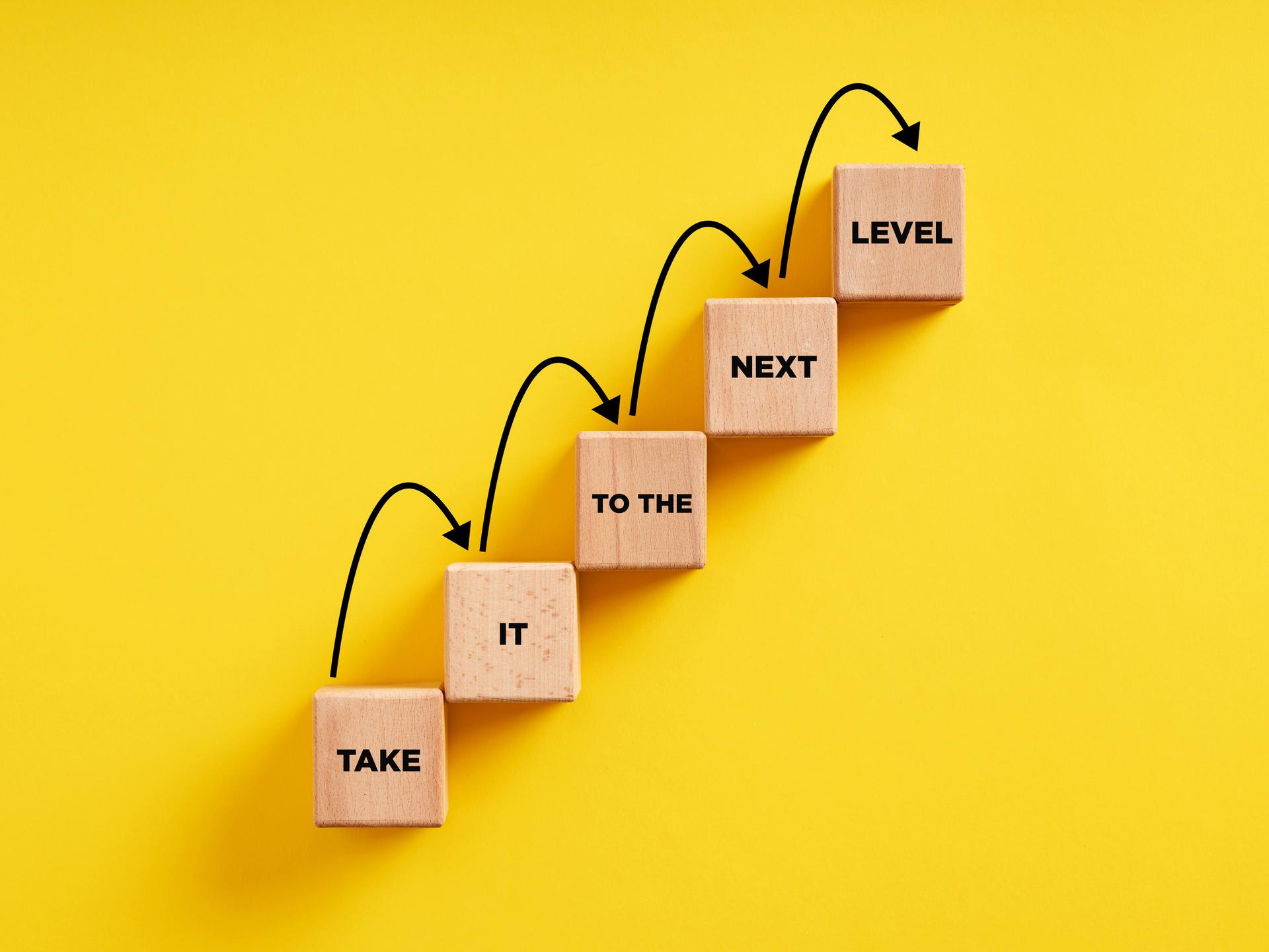 Business career path for growth and success concept. Wooden block ladder with the message take it to the next level with stepping up arrows.